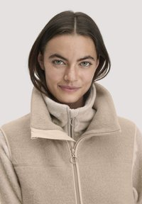 Beige fleece-takki, jossa on korkea kaulus, vetoketjukiinnitys ja kontrastiväri kauluksessa. Pehmeä tekstuuri ja toimiva muotoilu, jossa on pyöreä vetoketju.