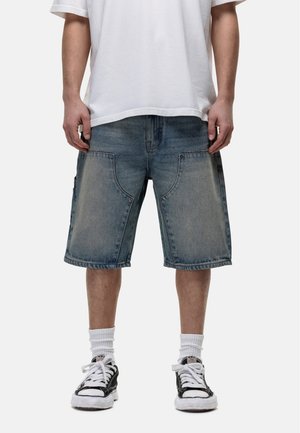 Denim shorts in light blue, loose fit with side pockets, finished with raw hems. Paired with a plain white t-shirt and black sneakers.