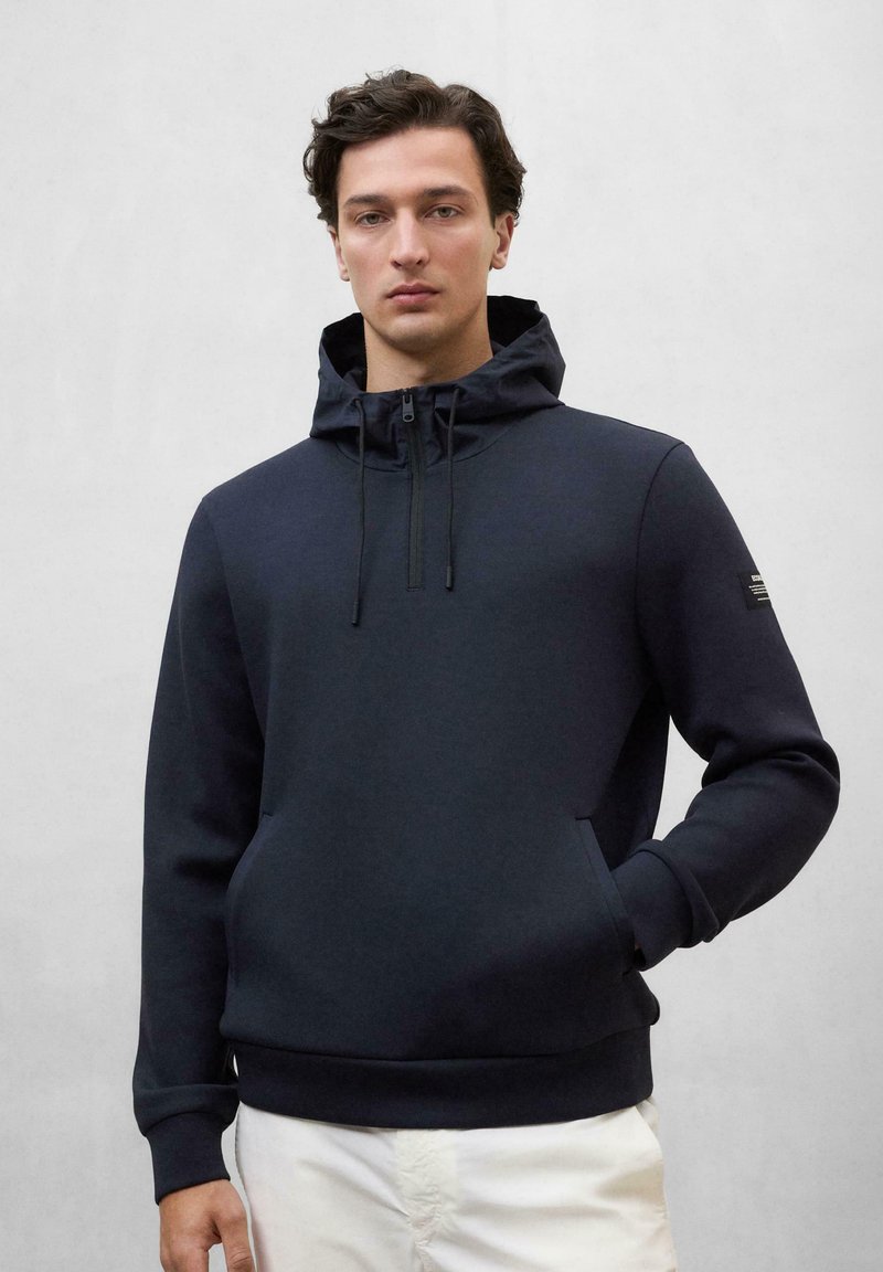 Sudadera con capucha de color navy, con cuello de cremallera, bolsillo tipo canguro frontal y puños elásticos. Presenta una etiqueta de marca en la manga izquierda.