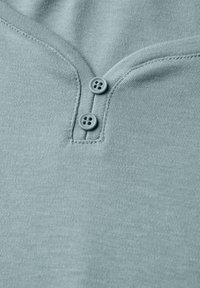 Himmelblaues Baumwoll-Henley-Shirt mit V-Ausschnitt, ausgestattet mit zwei runden, grauen Knöpfen am Kragen und einem weichen, strukturierten Stoff.