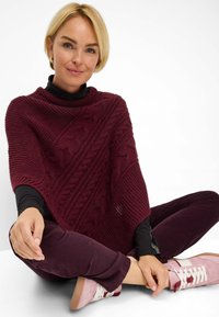 Burgunderfarbene Strickponcho mit einem strukturierten Kabelmuster, getragen über einem schwarzen Rollkragenpullover, kombiniert mit dunkelvioletten Hosen und hellrosa Sneakers.