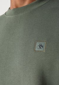 Oliven grøn crewneck sweatshirt med ribbet halsudskæring, prydet med et firkantet mærke med et broderet design i blå og beige på brystet.