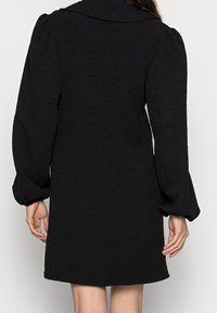 Kvinna i en texturerad svart långärmad klänning med puffärmar, sedd bakifrån mot en enfärgad ljus bakgrund.