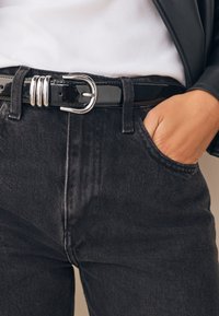 Jean en denim noir avec un lavage foncé, doté d'une ceinture à boucle argentée et de poches avant, présentant une texture lisse et une coupe ajustée.