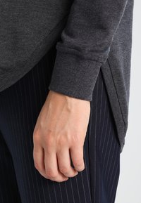 Haut gris à manches longues avec des poignets côtelés, présentant un design à fente sur le côté. Associé à un pantalon à rayures sombres. Texture de tissu lisse.