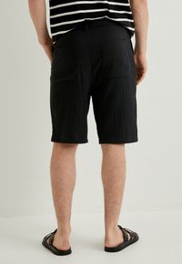 Sorte teksturerede shorts med to baglommer, der når til midten af låret. Båret med en sort og hvid stribet top og sandaler, stående op.
