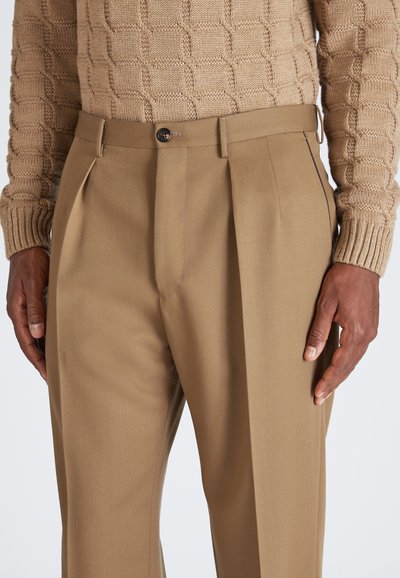 Beige maßgeschneiderte Hose mit vorderen Falten und Knopfverschluss. Glatter Stoff mit leichtem Glanz. Getragen mit einem strukturierten beige gestrickten Pullover.