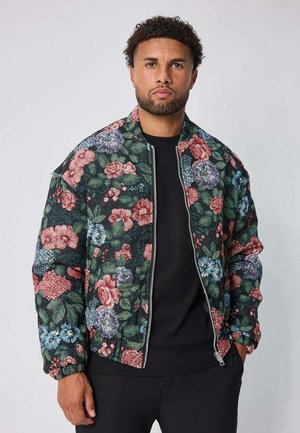 Homme aux cheveux bouclés portant une veste bomber zippée avec des motifs floraux multicolores sur un t-shirt noir, debout devant un fond uni.