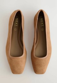 Tan ruskind slip-on sko med firkantet tå, der har en glat, bleg pink læderkant omkring åbningen og en polstret sål.