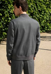 Dunkelgrauer Sweatshirt mit Kragen, strukturiertem Stoff, gerippten Bündchen und Saum. Kombiniert mit passenden Hosen, vor einem Hintergrund aus grünem Laub.