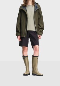 Personne portant une veste de pluie vert olive, un t-shirt beige, un short noir et de grandes bottes en caoutchouc vert olive sur un fond blanc.