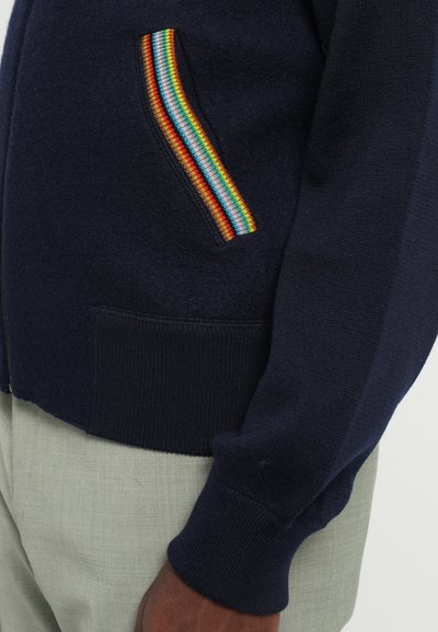 Marineblå stickad kofta med en flerfärgad randig fickdetalj och ribbade muddar. Har en slät textur och en figurnära design.