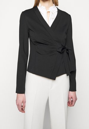 Nainen, joka pukeutunut mustaan käärittävään takkiin, jossa on sivusolmuke, valkoisen röyhelöidyn paidan päällä ja korkeavyötäröisiin valkoisiin housuihin, yksivärisen taustan edessä.