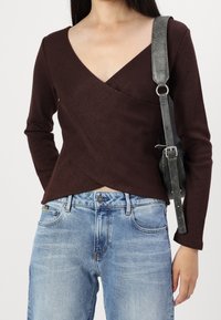 Brun långärmad omslagstopp med V-ringning, i kombination med blå denimjeans. En svart texturerad handväska bärs över axeln.