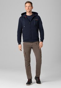 Navy zip-up hoodie featuring a kangaroo pocket and contrasting red drawstrings, paired with beige cargo pants and black lace-up boots.