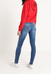 Sudadera recortada roja con letras negras, combinado con jeans ajustados azules. Zapatillas blancas visibles en los pies. La tela parece suave y elástica.