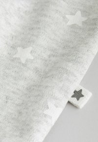 Grauer Stoff mit verstreuten hellgrauen Sternen und strukturierter Oberfläche. Am Rand ist ein kleines Stoffetikett mit einem dunkelgrauen Stern zu sehen.