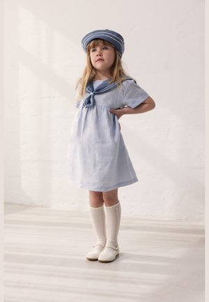 Νεαρή κοπέλα που φοράει μπλε φόρεμα ναυτικού και ταιριαστό καπέλο, λευκές κάλτσες μέχρι το γόνατο και λευκά παπούτσια, στέκεται με το ένα χέρι στη μέση.