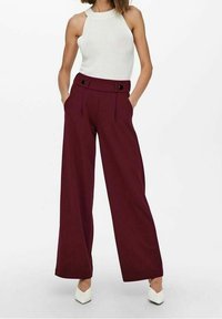 Bílý žebrovaný top na zavazování za krk v kombinaci s vysokými, širokými kalhotami v barvě burgundy, které mají dva černé knoflíky. Modelka stojí s rukama v kapsách.