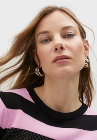 Orecchini a cerchio dorati con lettering nero, che recano la parola "AMORE". La modella indossa un maglione a righe nere e rosa con collo rotondo.