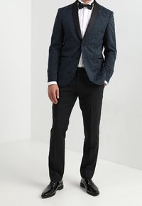 Uomo che indossa un blazer a fantasia blu scuro, camicia bianca, papillon nero, pantaloni neri e scarpe da cerimonia nere lucidate, in piedi all'interno.