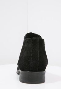 Black suede ankle boot with a smooth rounded toe, textured upper, and a low black stacked heel. Features visible stitching along the seams.