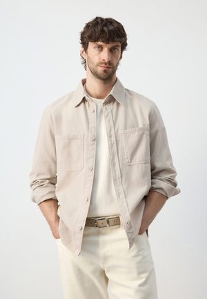 Moški s kratkimi kodrastimi lasmi in brado, ki nosi bež srajco z gumbi, belo majico, kremne hlače in bež pas z rokami v žepih.