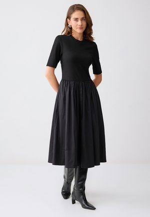 Femme portant une robe midi noire à manches courtes et jupe froncée, associée à des bottes à talons noires montantes jusqu'aux genoux et des boucles d'oreilles décoratives.