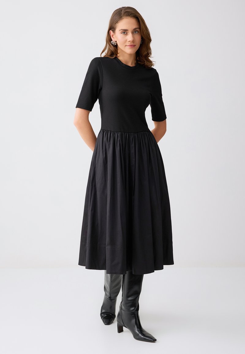 Femme portant une robe midi noire à manches courtes et jupe froncée, associée à des bottes à talons noires montantes jusqu'aux genoux et des boucles d'oreilles décoratives.