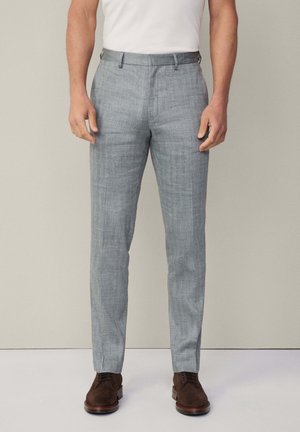 Pantalons taillés gris clair en tissu tissé, avec une coupe plate, des passants de ceinture et une texture subtile. Associés à des chaussures marron.