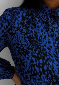 Blaue Bluse mit einem Leopardenmuster in Schwarz. Knöpfe entlang der Vorderseite und geraffte Ärmel verleihen Textur und Detail.