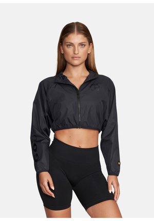 Kobieta ubrana w czarną, krótką kurtkę z zamkiem i elastycznym dołem oraz czarne, dopasowane szorty z wysokim stanem, stojąca na tle jasnego, jednolitego tła.