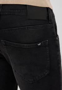 Los jeans de mezclilla negra presentan un acabado texturizado, con costuras visibles, una etiqueta de cuero en la parte posterior y un pequeño logo de caballo en el bolsillo.