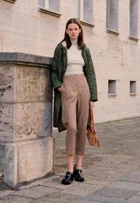 Cappotto verde trapuntato sopra un leggero dolcevita, abbinato a pantaloni a quadri marroni e scarpe con tacco plateau nere. Tiene in mano una borsa a tracolla marrone.