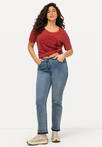 Camiseta roja casual con detalle anudado, que presenta un gráfico circular. Jeans de cintura alta de color azul claro con un leve estiramiento, de longitud hasta los tobillos. Zapatillas de color crema.