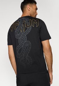Svart bomullst-skjorte med et stort, teksturert slangemotiv i grått og "VENUM"-bokstaver i gull på baksiden. Crew neck-design.