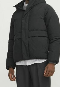 Schwarze Steppjacke mit Kapuze, mit gesteppter Musterung und elastischen Bündchen. Wird über einem hellgrauen Pullover und einem gestreiften Hemd getragen.