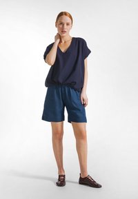 Frau mit roten Haaren, die ein lockeres marineblaues V-Ausschnitt-Top, Jeansshorts und burgunderrote Schnürschuhe trägt und vor einem weißen Hintergrund steht.