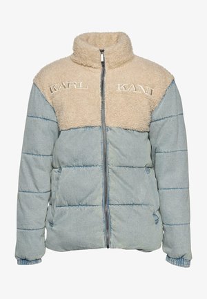 Chaqueta acolchada azul claro con paneles de forro polar beige en el pecho bordados con "KARL KANI", llevada sobre una camisa blanca, manos cruzadas mostrando tatuajes y anillos.