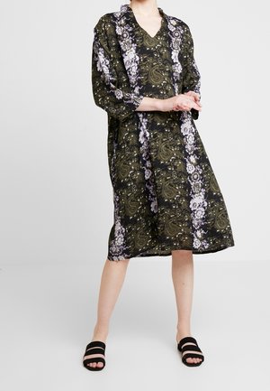 Kvinde iført en løs, knælængde mørk kjole med grøn paisley og hvide blomsterprint, og sorte sandaler med to remme, stående med hænderne foldet.