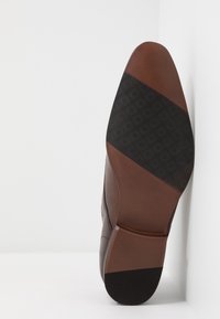 Hnědé kožené boty s texturovanou černou gumovou podrážkou. Mají hladký svršek se geometrickými vzory a kontrastními akcenty na špičce a patě.