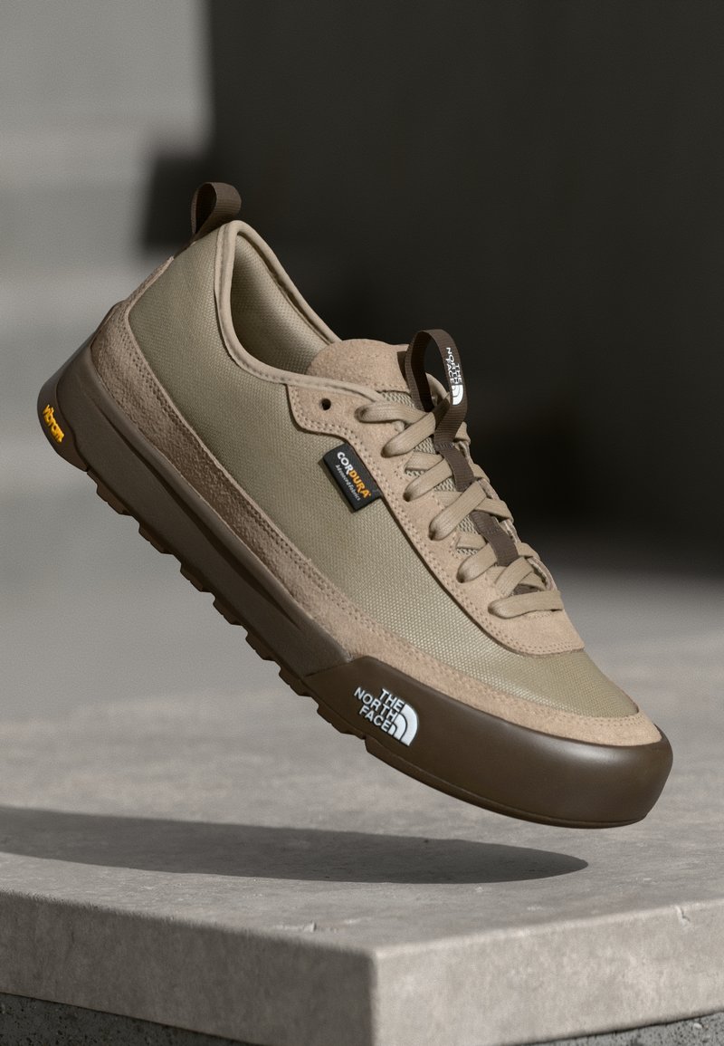Beige athletic shoe with mesh and suede upper, brown rubber sole, cushioned, featuring logo accents and lace-up design.