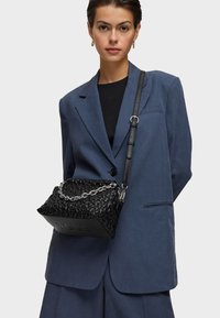 Giovane donna che indossa un completo azzurro oversize e un top nero, mentre porta una borsa nera strutturata con una tracolla a catena argento sulla spalla.