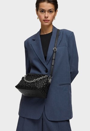 Giovane donna che indossa un completo azzurro oversize e un top nero, mentre porta una borsa nera strutturata con una tracolla a catena argento sulla spalla.