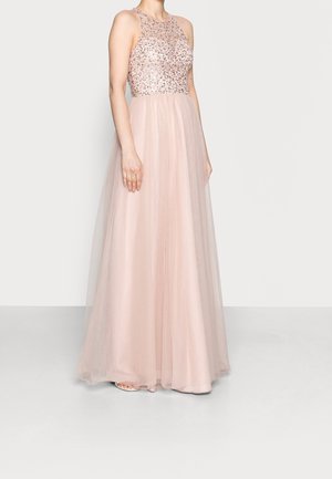 Femme portant une robe sans manches rose pâle avec un corsage scintillant et orné et une jupe en tulle fluide, debout devant un fond uni.