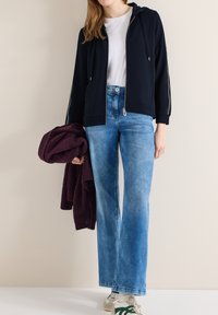 Personne portant un sweat à capuche zippé bleu marine, un t-shirt blanc, un jean bleu clair et des baskets blanches, tenant une veste pliée de couleur violet foncé à côté d'elle.