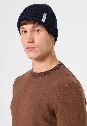 Junger Mann trägt eine marineblaue Strickmütze mit Etikett und einen braunen Rundhalspullover, schaut leicht zur Seite vor einem einfarbigen Hintergrund.