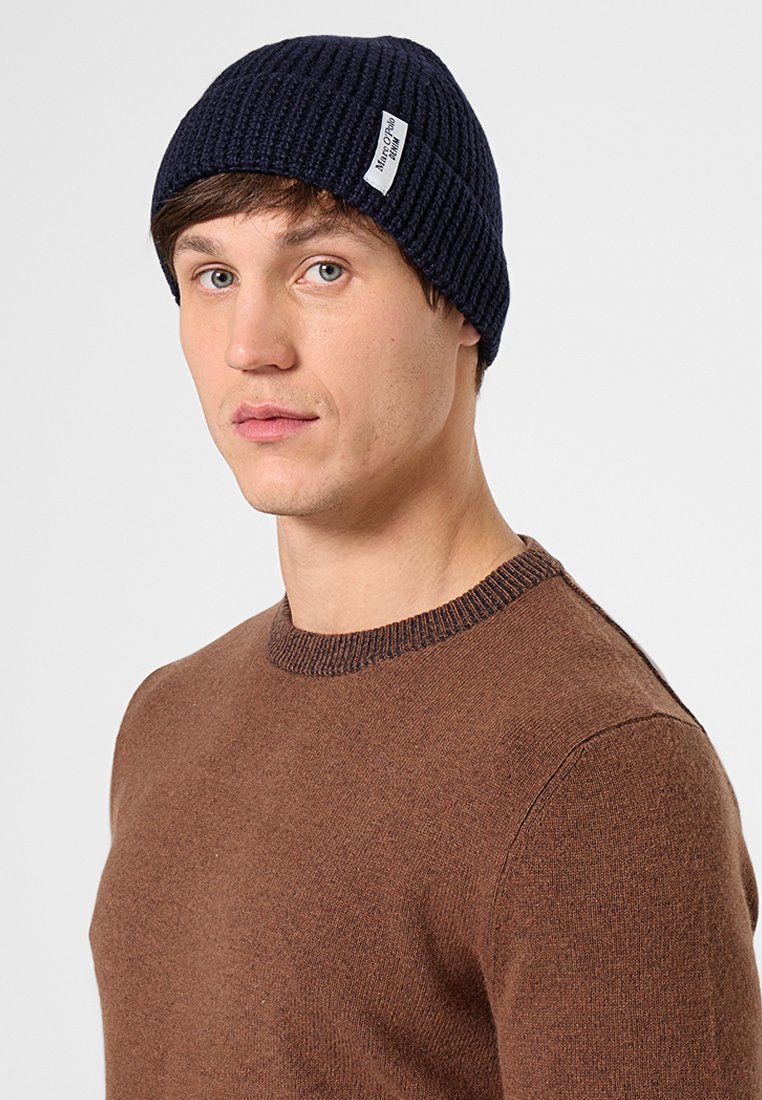 Young man wearing a navy knit beanie with label and a brown crewneck sweater, looking slightly to the side against a plain background.