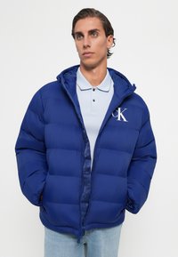 Ung mand iført en blå puffjakke med CK-logo over en lys blå polo skjorte og lyseblå jeans, stående foran en ensfarvet baggrund.