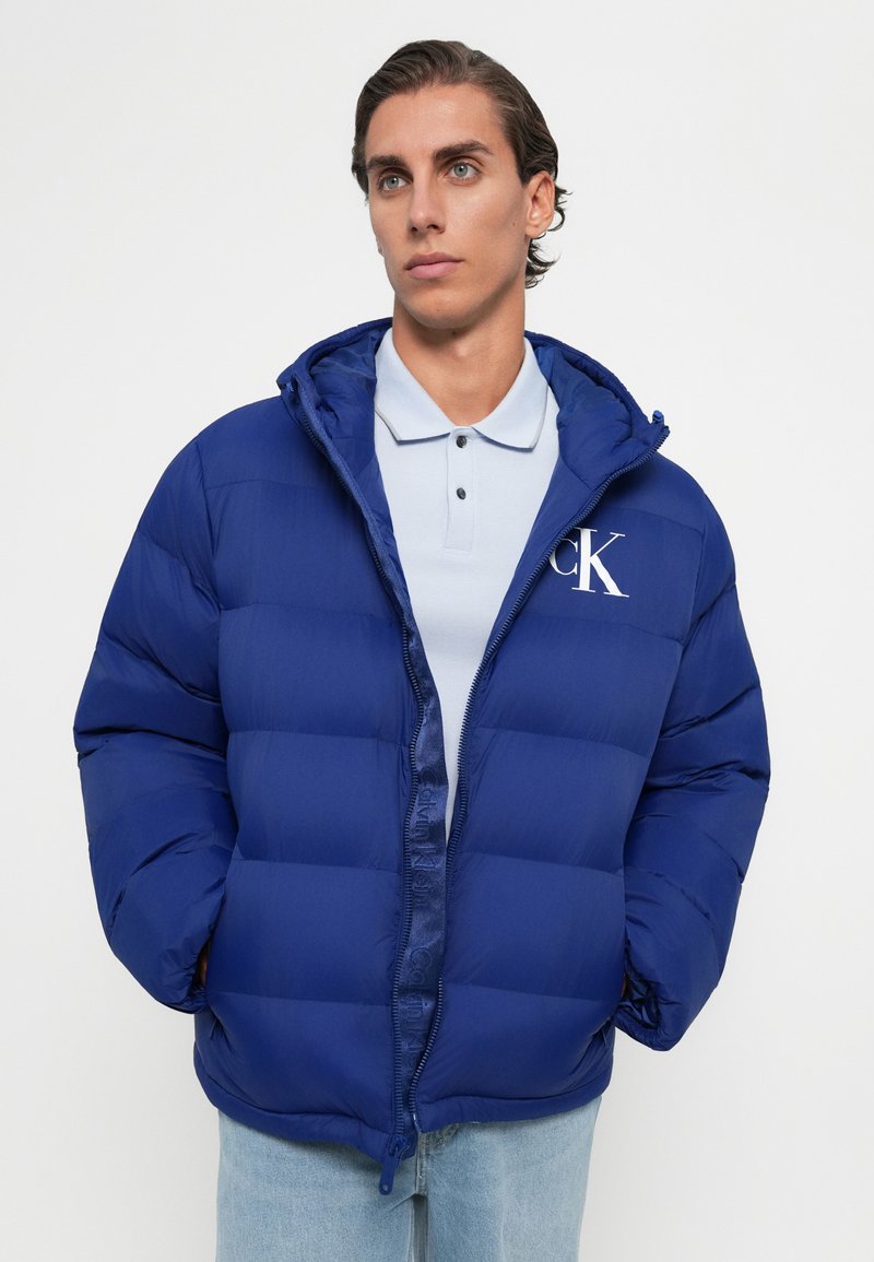 Fiatal férfi kék pehelykabátot visel CK logóval egy világoskék póló és világoskék farmer mellett, egy sima háttér előtt áll.
